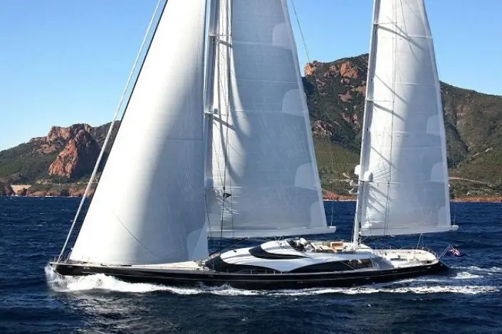 Crewed sailing yacht under full sail along a rocky Mediterranean coastline